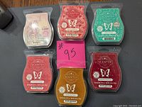 Photo showing 6 Scentsy scent wax bars arranged in two rows on a gray surface with a pink #95 tag. Each wax bar is sealed and displays distinct label colors and designs.
