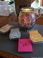 Glass Scentsy wax warmer with colorful firework design and three different partially used Scentsy wax melt sets placed around it.