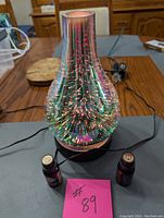 Full front view of the glass electric diffuser, showing its reflective, colorful surface, and two small oil scent bottles placed in front with a pink tag #89.