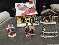 Close-up photo of yellow cabin with snow-covered roof and three figurines: two carolers and a family group, along with a small fountain figurine.