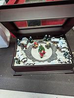 Wooden music box opened showing detailed winter village scene with Santa Claus figure in center, snow-covered trees and houses on a cobblestone base inside a dark wood box with glass lid.