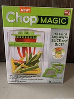 Front view of ChopMAGIC box showing product image with sliced vegetables inside the container, branding, features, and included blades.