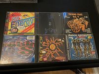 Front view of six vintage music CD cases arranged on a flat dark surface. Covers show colorful, energetic designs with text and graphics representing various dance and techno music compilations.