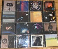 Photo shows a collection of 16 sealed Kitaro music CDs arranged closely in four rows of 4. Each jewel case has distinctive album art with various images and colors including abstract designs, landscape, and spiritual themes.