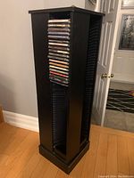 View of one side of the tall black wooden CD holder showing vertical rows of CDs in upright position, highlighting the storage capability and condition with some visible scratches.