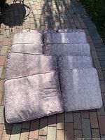 Seven light purple patio chair cushions, stacked and laid out on a brick patio surface, showing their floral patterns and slight discoloration.