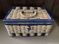 Closed wicker sewing organizer box with blue trim and latch, woven handle on lid, on wooden surface.