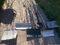 Top down view of six snow shovels laid on brick pavement showing both metal and plastic blades and wooden and metal handles.