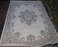 Image showing full view of a light beige wool carpet with floral patterns and fringed edges laid outdoors on pavement and grass.