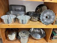 Photo of two bundt pans, two rectangular roasting pans with lids on an upper shelf, two large stewing pots with lids and one large metal mixing bowl on a lower shelf.