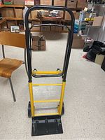 Front view of the yellow hand truck dolly standing upright with black handle and base plate, visible in a basement room with miscellaneous storage boxes and furniture.