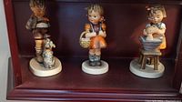 Three Goebel Hummel porcelain figurines displayed on a shelf, showing their relative sizes and colors.
