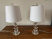 Pair of clear resin table lamps on wooden surface, showing white plastic shades and clear geometric bases.