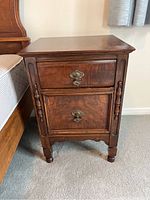 Front view of vintage wooden nightstand showing two drawers with ornate metal handles and carved wood details on legs and apron.