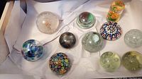 Top-down view of a box containing 11 assorted glass and acrylic paperweights of various shapes and colors.