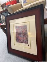 Framed print in its entirety showing the dark wood frame with a red inner border and cream matting around the print.