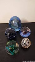 All five glass paperweights displayed on a black surface showing various colors and sizes, the largest about 4 inches tall.
