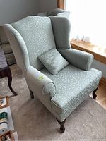 Front-left 3/4 view of wingback armchair with decorative cushion on seat, showing wood cabriole legs and overall upholstery pattern