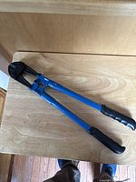 Bolt cutter shown from above on wooden surface, blue handles with black grips and black cutting head visible.
