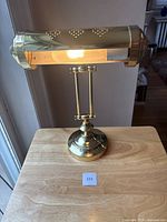 Full view of the brass desk lamp turned on, showing the light illuminating inside the cylindrical brass shade with perforations.