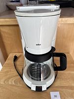 Front view of Braun white coffee maker with glass carafe and black handle, power cord visible.