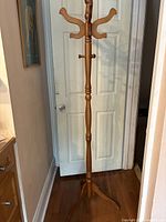 Front view of the full-height wood coat rack standing in front of a white door, showing the smooth wooden pole and upper hooks.