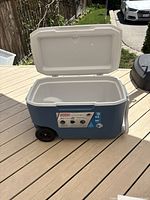 Side view of cooler with lid open showing blue body, white lid, two black wheels and handle at side.