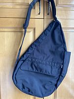 Front view of black Ameri Bag cross body travel organizer hanging on wooden door, showing overall teardrop shape with flap pocket on front and padded adjustable strap.