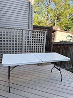 Full view of folding table set up outdoors on a deck, showing white plastic top and black metal legs.