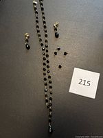 Full view of gold chain necklace with black crystal beads, black teardrop earrings, and 4 loose black crystal bead dangles arranged on dark background with lot number card.