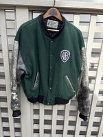 Front view of the Roots 1995 jacket showing green fabric body, worn grey sleeves, and Warner Bros. logo patch on left chest area.