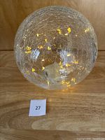 Clear spherical crackle glass globe illuminated by yellow twinkle lights inside, placed on wooden surface with a number 27 label.