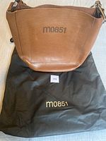 Front view of the M0851 cognac leather bucket style shoulder bag, clearly showing embossed M0851 logo and included black branded storage bag underneath.