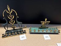 Two brass and enamel menorahs placed side by side on a table with black background. Left menorah features two deer and a leafy branch design, right menorah has a soldier figure and green speckled enamel base with candle holders.