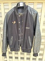 Front view of men's black wool jacket with leather sleeves, showing button closure, ribbed collar, and embroidered Adderly logo with airplane patch.