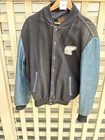 Front view of black wool crew jacket with blue leather sleeves, snap buttons, and bear patch on chest.