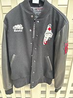 Front view of the black varsity style jacket showing embroidered white Roots logo on right chest and stylized face logo on left chest along with button snap closure and leather sleeves.