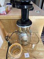 Omega VRT 350 juicer main unit with attachments, placed on wooden surface. Includes clear plastic juice and pulp containers and small orange-colored juicer part still in plastic wrap.