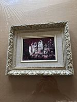 Framed oil painting showing a city nightscape with illuminated buildings, people, and umbrellas, in an ornate cream frame, front view.