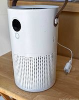 Front view of white cylindrical Proton Pure air purifier with button, sensor, brown handle, and power plug visible on wooden surface.