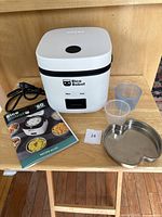 Rice Robot cooker, power cord, two measuring cups, stainless steel steamer tray, recipe book, and a serving spoon on a wooden surface