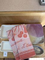Two small white textured rectangular platters with handles, one larger (11x6) and one smaller (8x4), positioned side-by-side with a multicolor pottery garlic set and two folded peach and white tea towels placed on top.
