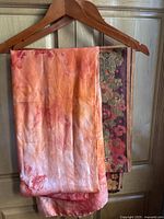 Two silk scarves hanging on wooden hangers against a wooden door, showing the orange tie-dyed scarf in front and the multicolor floral woven scarf behind.