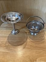 Both sterling silver pierced candy bowls shown side by side on a wooden surface, one taller pedestal style and one smaller handled bowl.