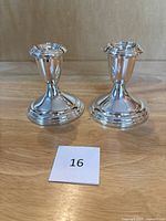 Front view of the pair of polished silver-toned sterling weighted candlesticks, placed on a wooden surface with a label "16" in front.