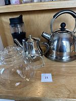 Lot showing all items: silver stainless steel stove top kettle, silver small tea pot, clear glass tea pot, and clear Starbucks travel press with black silicone grip.