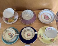 Full view of 5 teacups and saucers on wooden surface, showing floral motifs and varied color backgrounds including blue, yellow, and iridescent pink