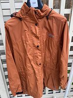 Rust-colored water-resistant MEC jacket, size 8, hanging on rack showing front zipper and snap closure with adjustable hood