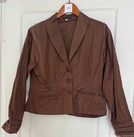 Front view of shiny chocolate brown silk shirt/jacket on hanger, showing 3 buttons and pocket details.
