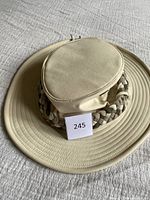 Front view of beige Tilley hat with oval crown, decorative braid band, and adjustable chin strap laid flat on bed.